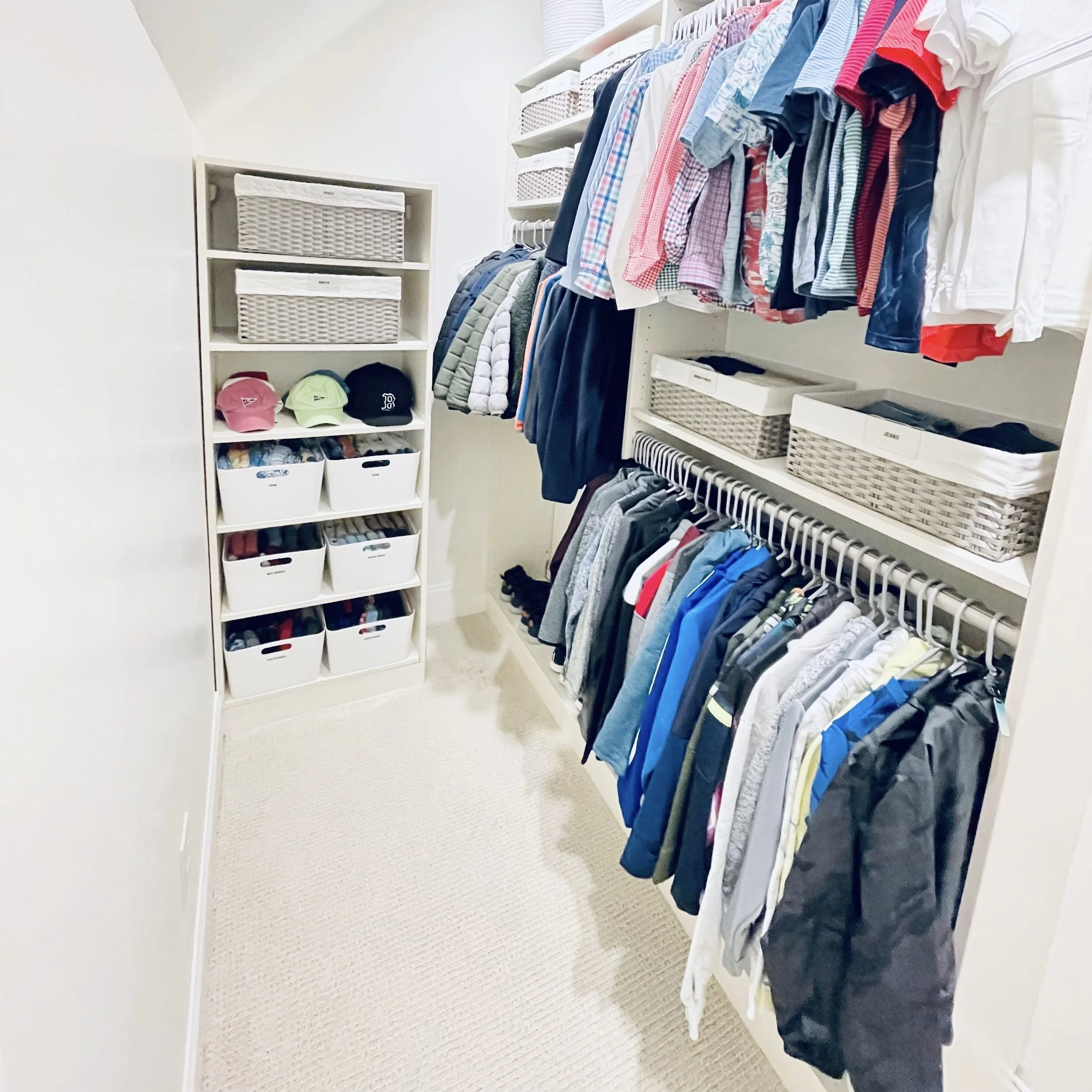 A long narrow walk-in closet with baseball caps organized on shelving with white handled bins and baskets, racks of clothes on the wall decluttered by Sorted Professional Organizing in New Orleans.