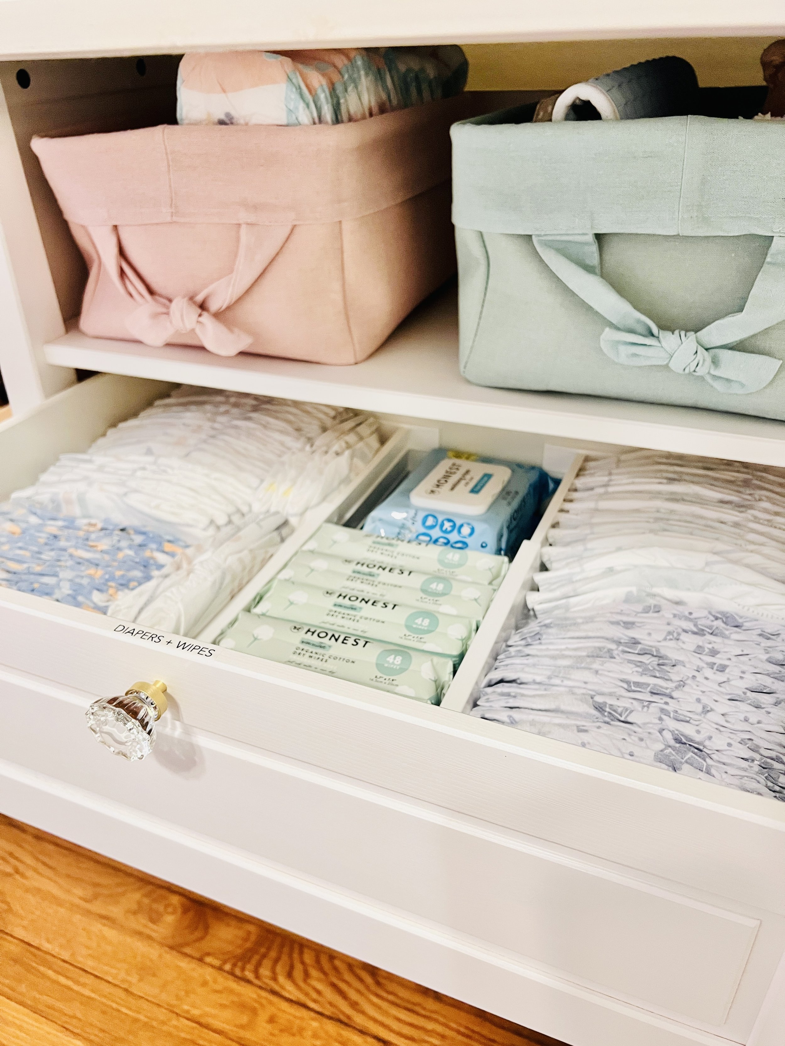 Open white drawer with labeled section holding diapers and wipes, containing folded cloths, tissue boxes, and fabric bins on a shelf above.