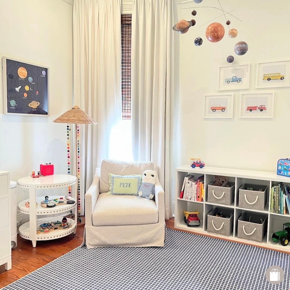 Tidied playroom with an armchair, colorful floor lamp, tiered end table, cubbies of organized books, toy bins, framed truck art, and a mobile solar system by Sorted Professional Organizing in New Orleans.