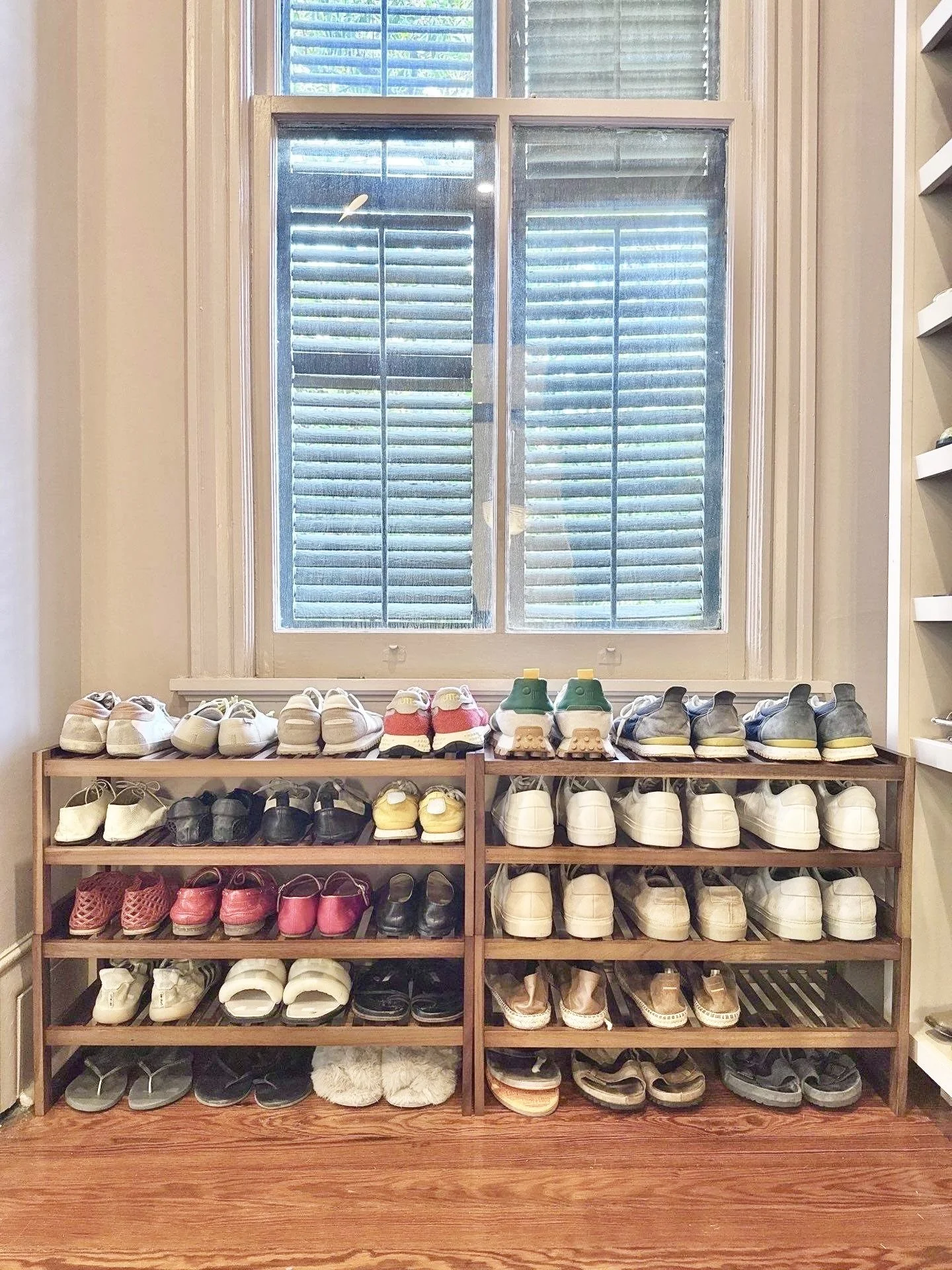Two show shelves of four shelves each with shoes organized on them against a wall underneath a window with sunlight coming through a dark louvered shutter.
