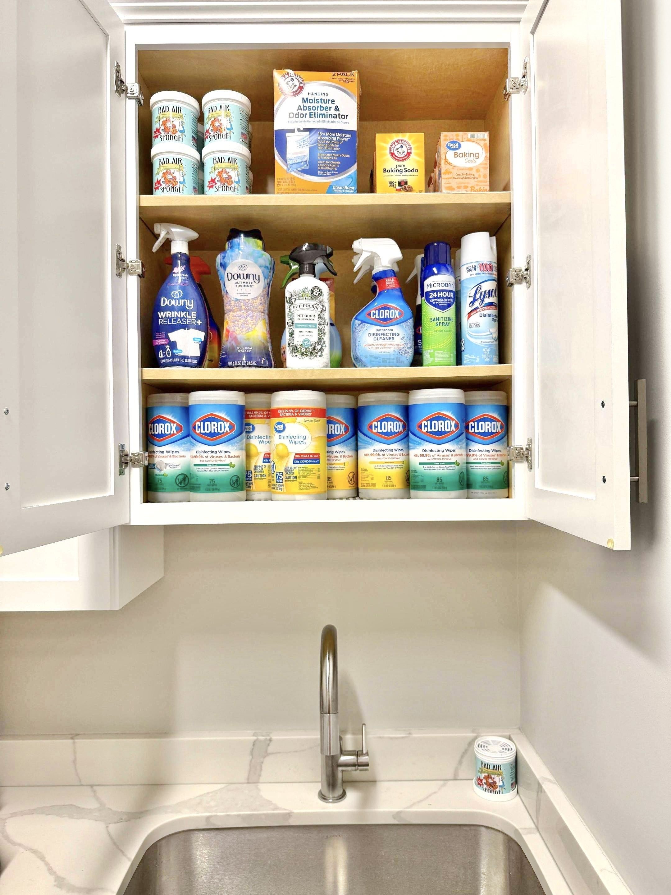 Utility room cabinet above a sink with neatly organized Clorox wipes, Downy products, stain removers, Lysol spray, and other cleaning products.