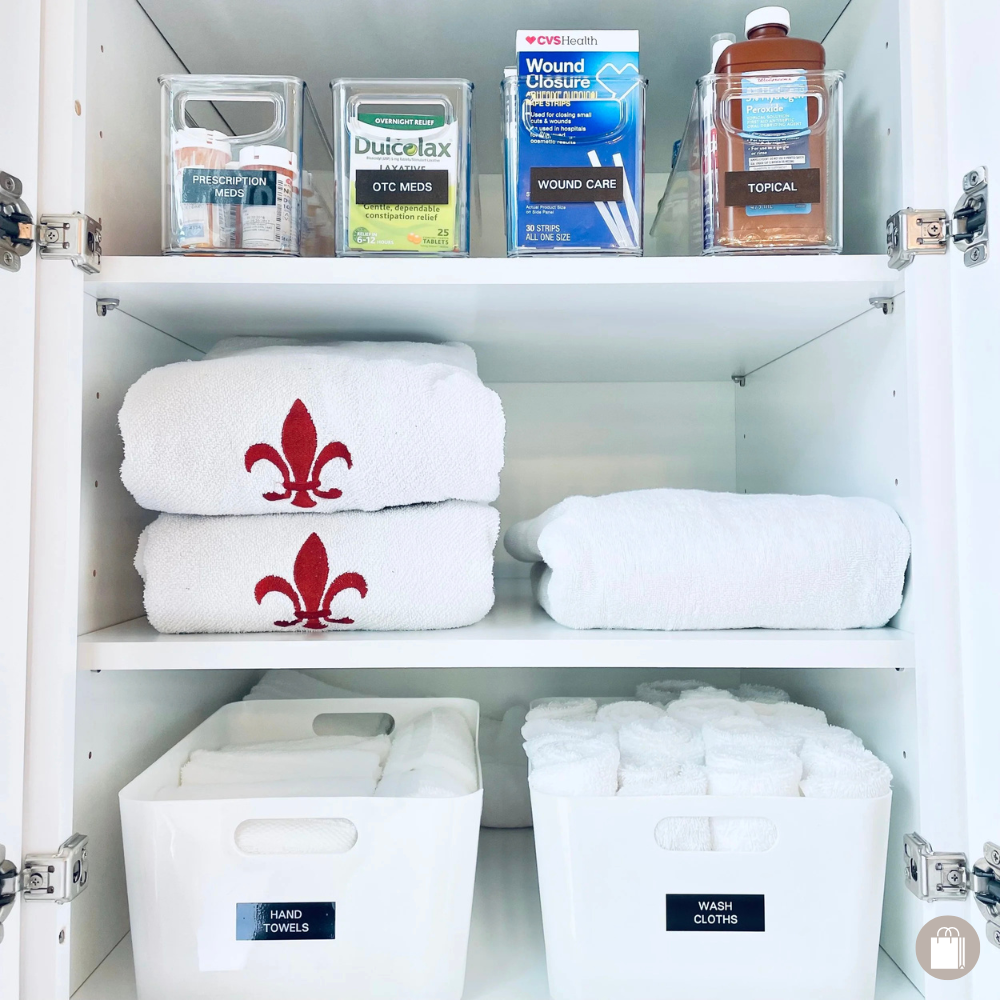 Linen closet cabinet with white-handled bins holding hand towels, neatly folded bath towels, and The Home Edit labeled acrylic bins for first aid products, carefully arranged by Sorted Professional Organizing in New Orleans.