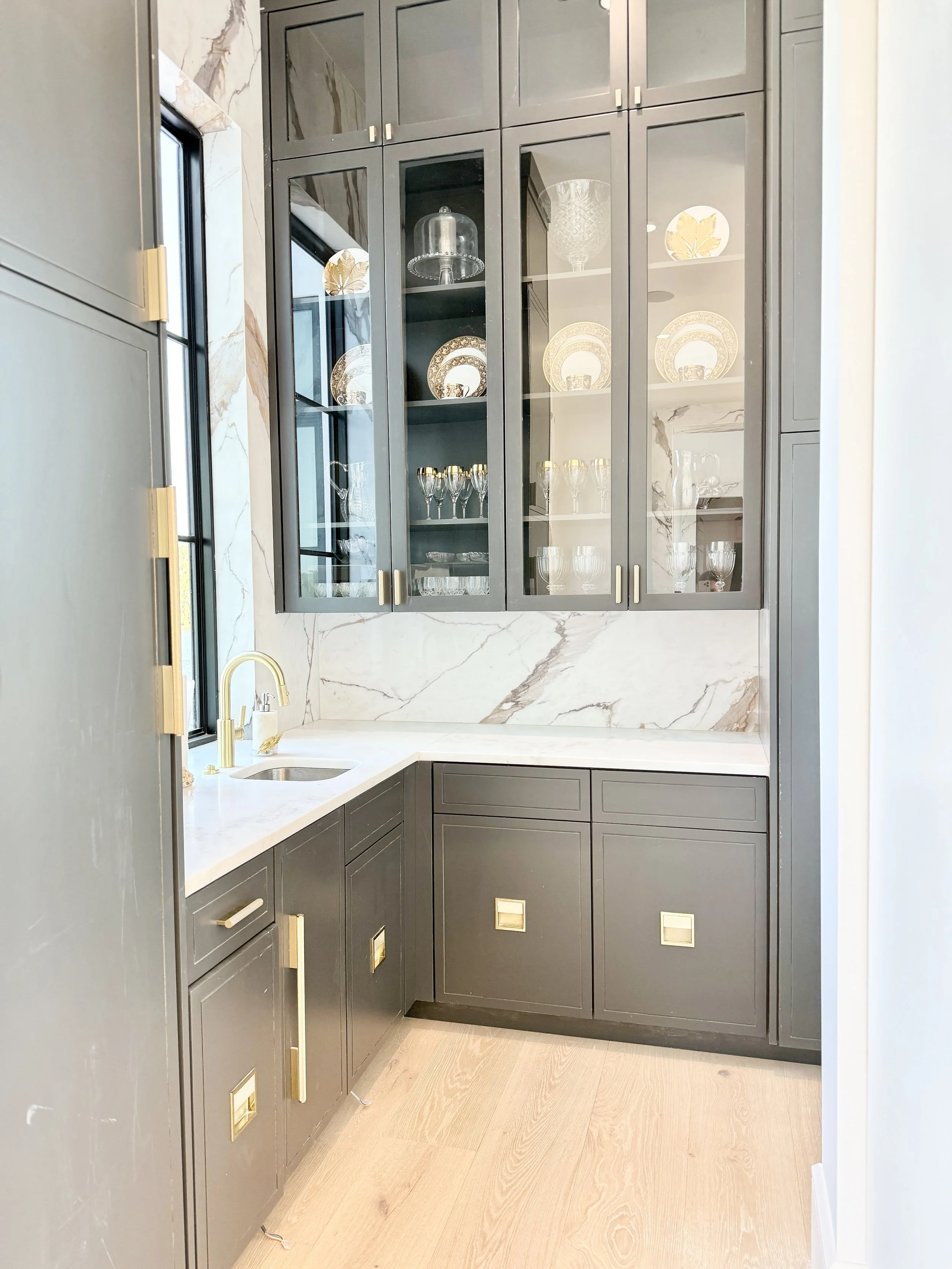 A modern kitchen corner with gray cabinets, a white marble countertop, and a matching marble backsplash. The upper glass-front cabinets display glassware and decorative plates. There is a small stainless steel sink with a gold faucet below a window, and the floor is light wood.
