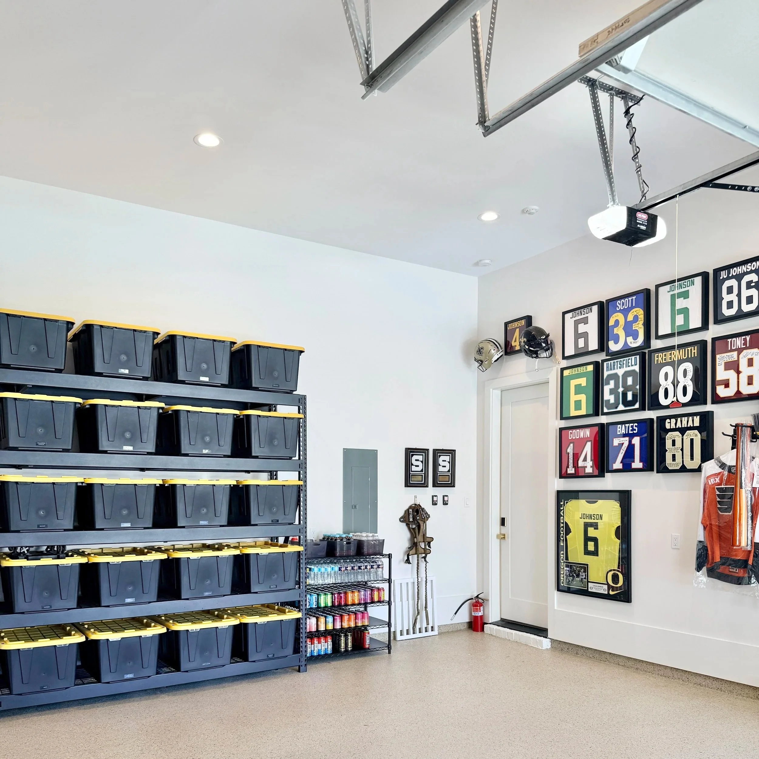 A sports memorabilia room with framed jerseys and helmets on the wall, black storage bins on metal shelves, and shelves with spray paint and sports equipment.