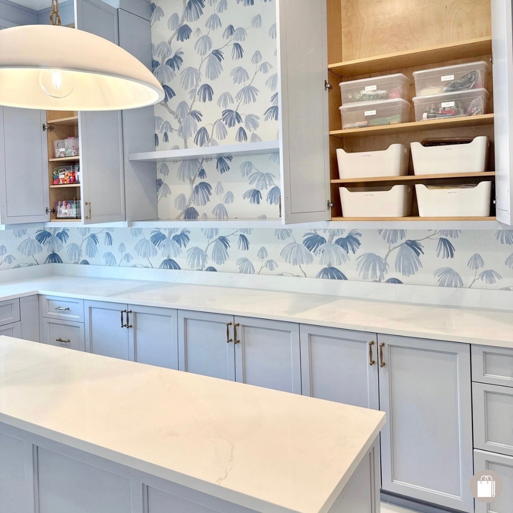 Beautiful laundry room with blue floral wallpaper, clear countertops, clean island, open cabinets with white bins, and clear labeled containers.