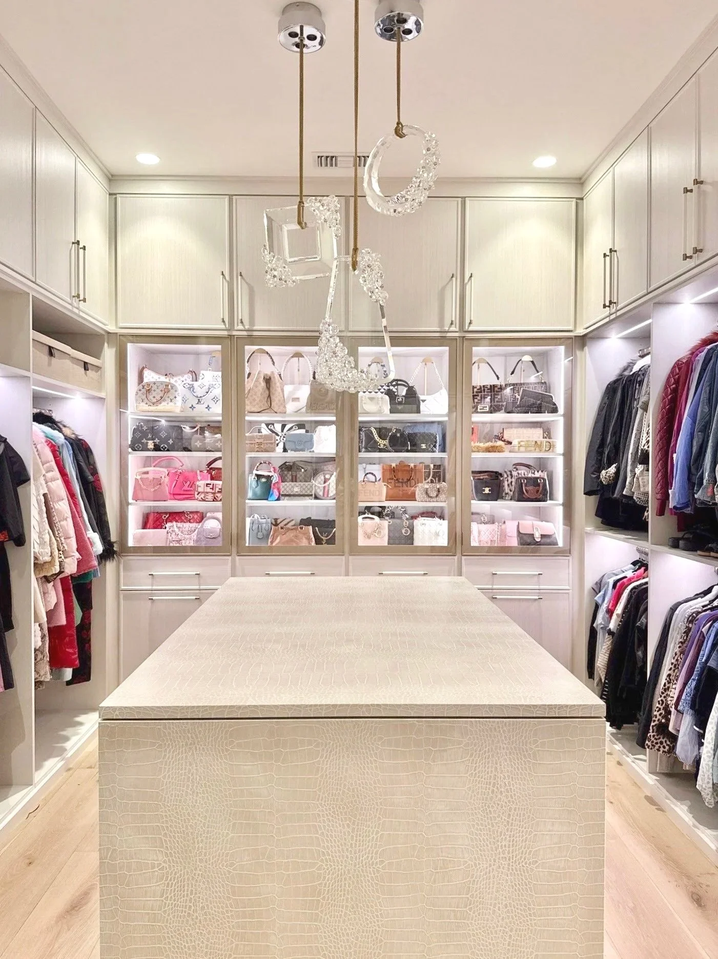 A walk-in closet with white built-in shelves storing handbags and clothes, a beige patterned island in the center, and crystal chandelier lighting.