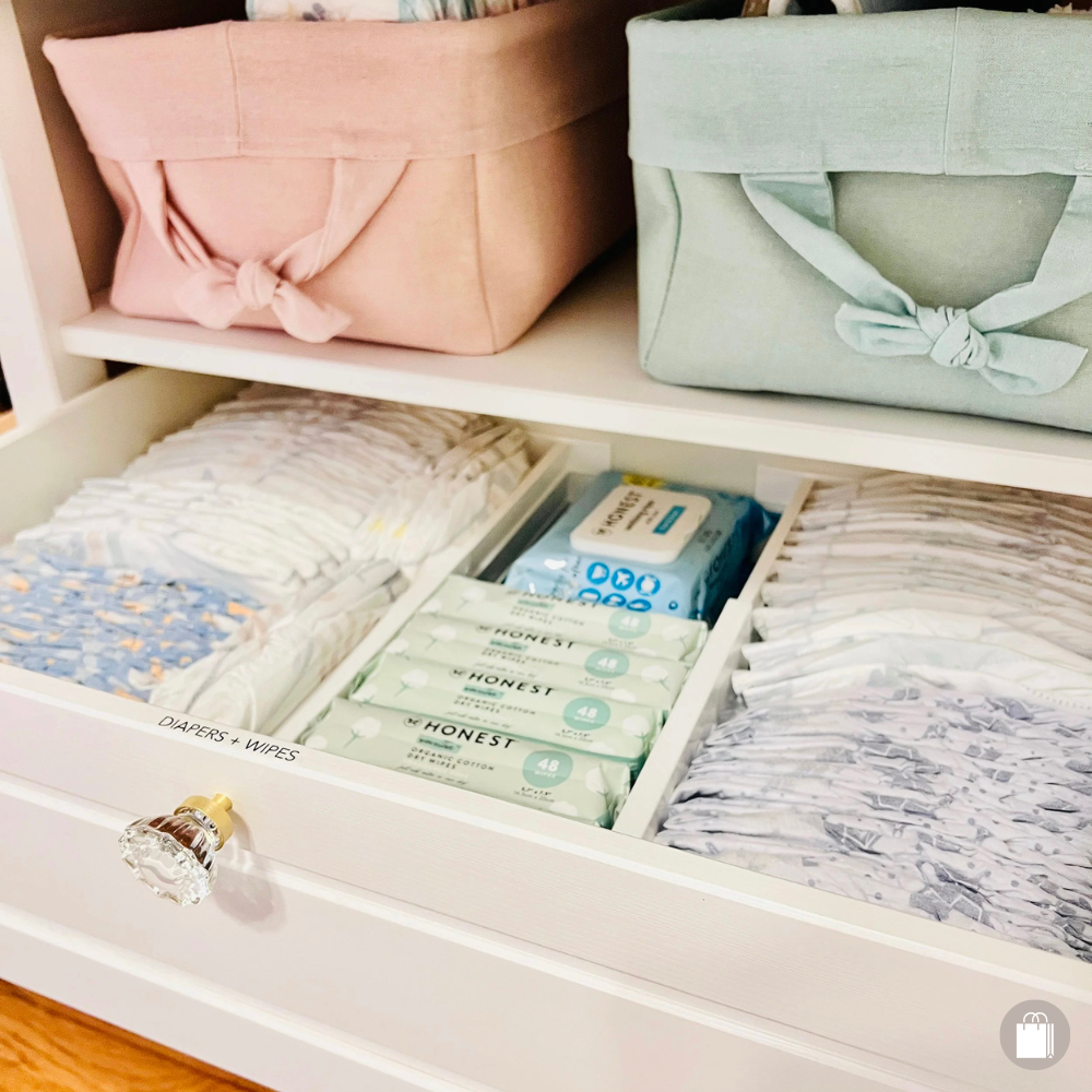 Open white drawer with labeled section holding diapers and wipes, containing folded cloths, tissue boxes, and fabric bins on a shelf above.