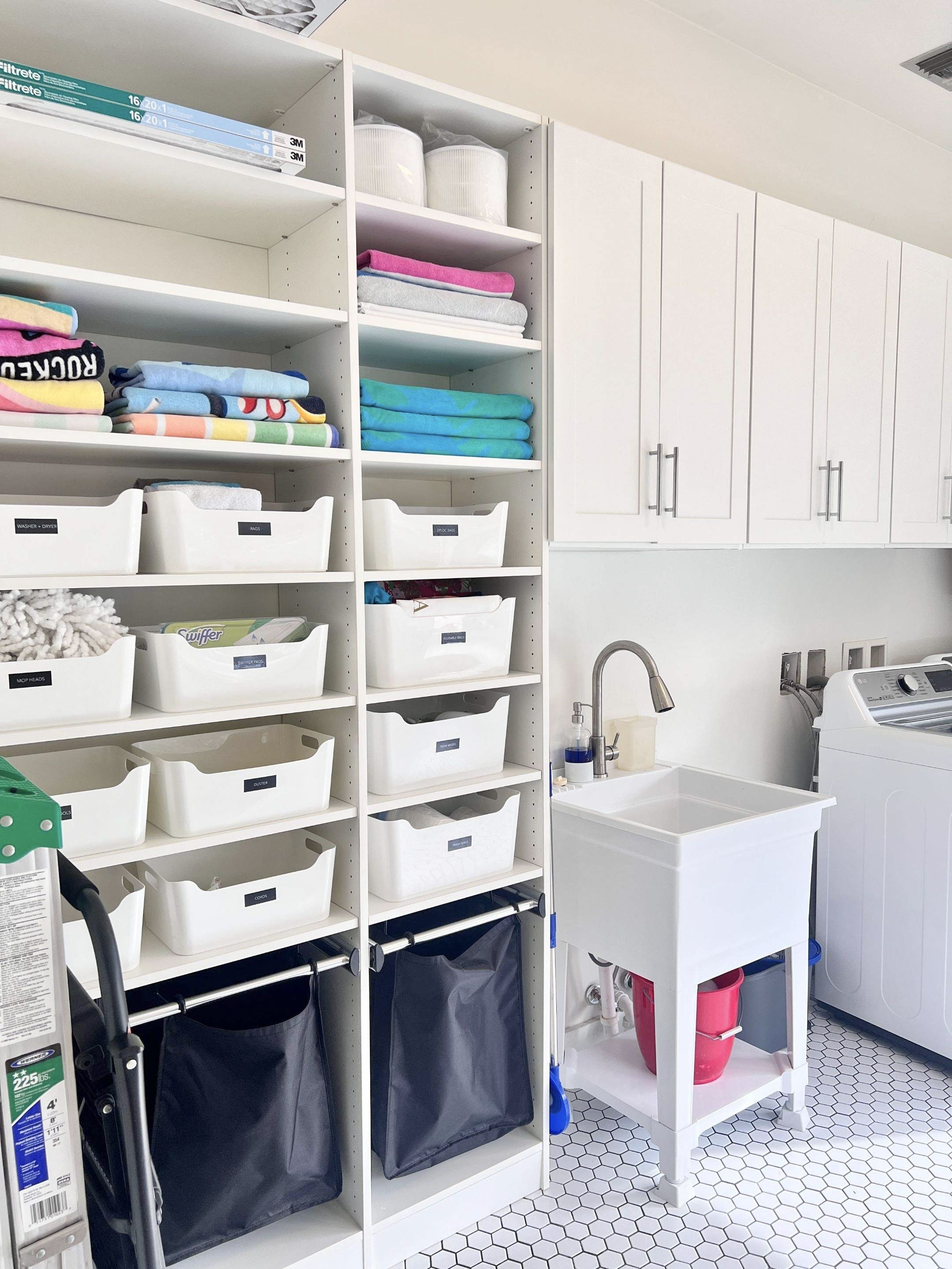 Laundry room with shelves holding folded towels, storage bins, and cleaning supplies, with a laundry sink and washing machine.