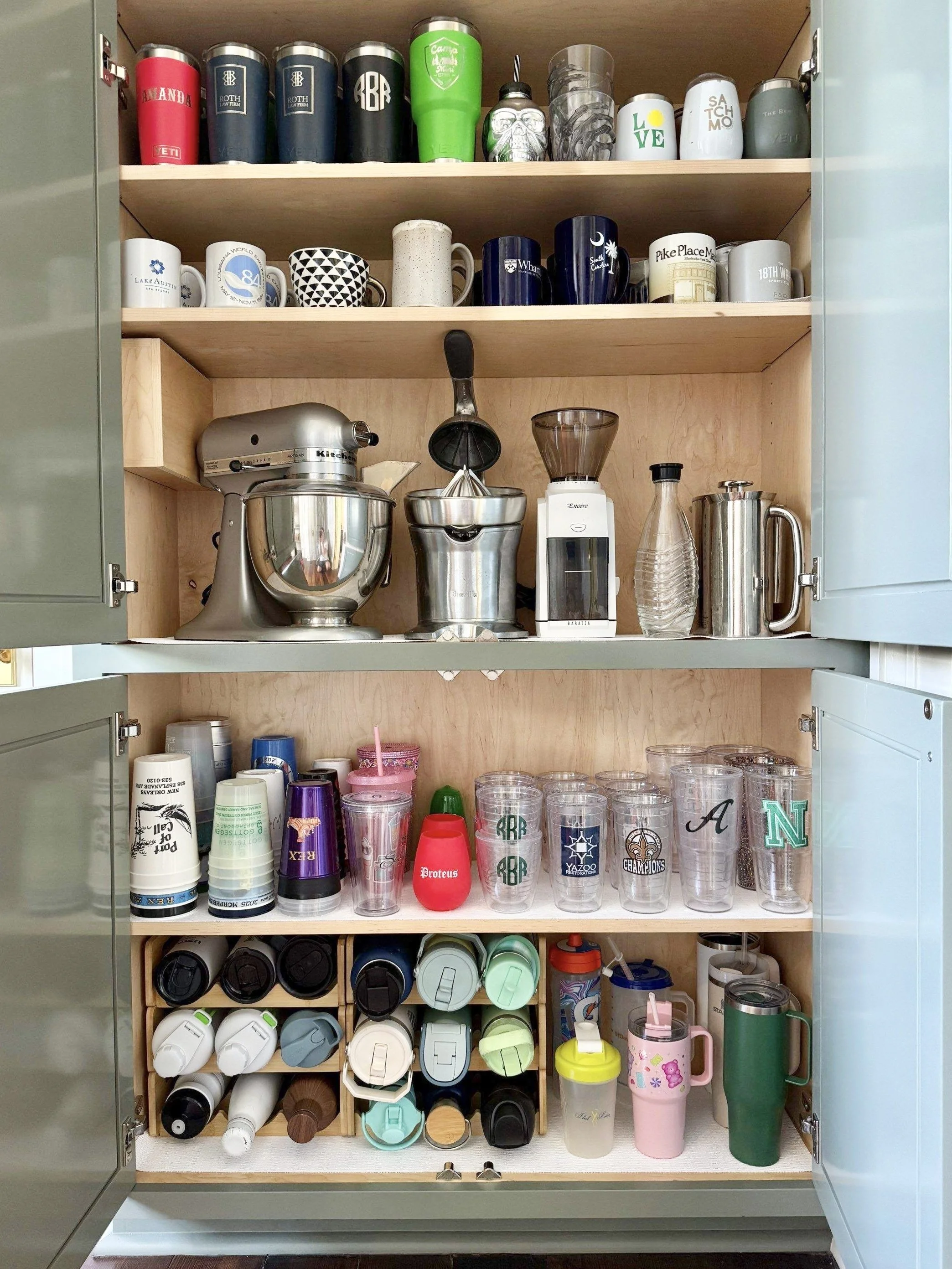 Sage green kitchen cabinet with neatly organized water bottles, tumblers, coffee mugs, and small appliances such as a mixer, juicer, and coffee maker.