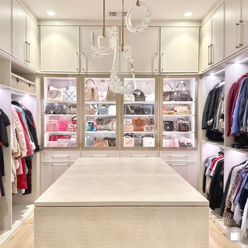 A walk-in closet with white built-in shelves storing handbags and clothes, a beige patterned island in the center, and crystal chandelier lighting.