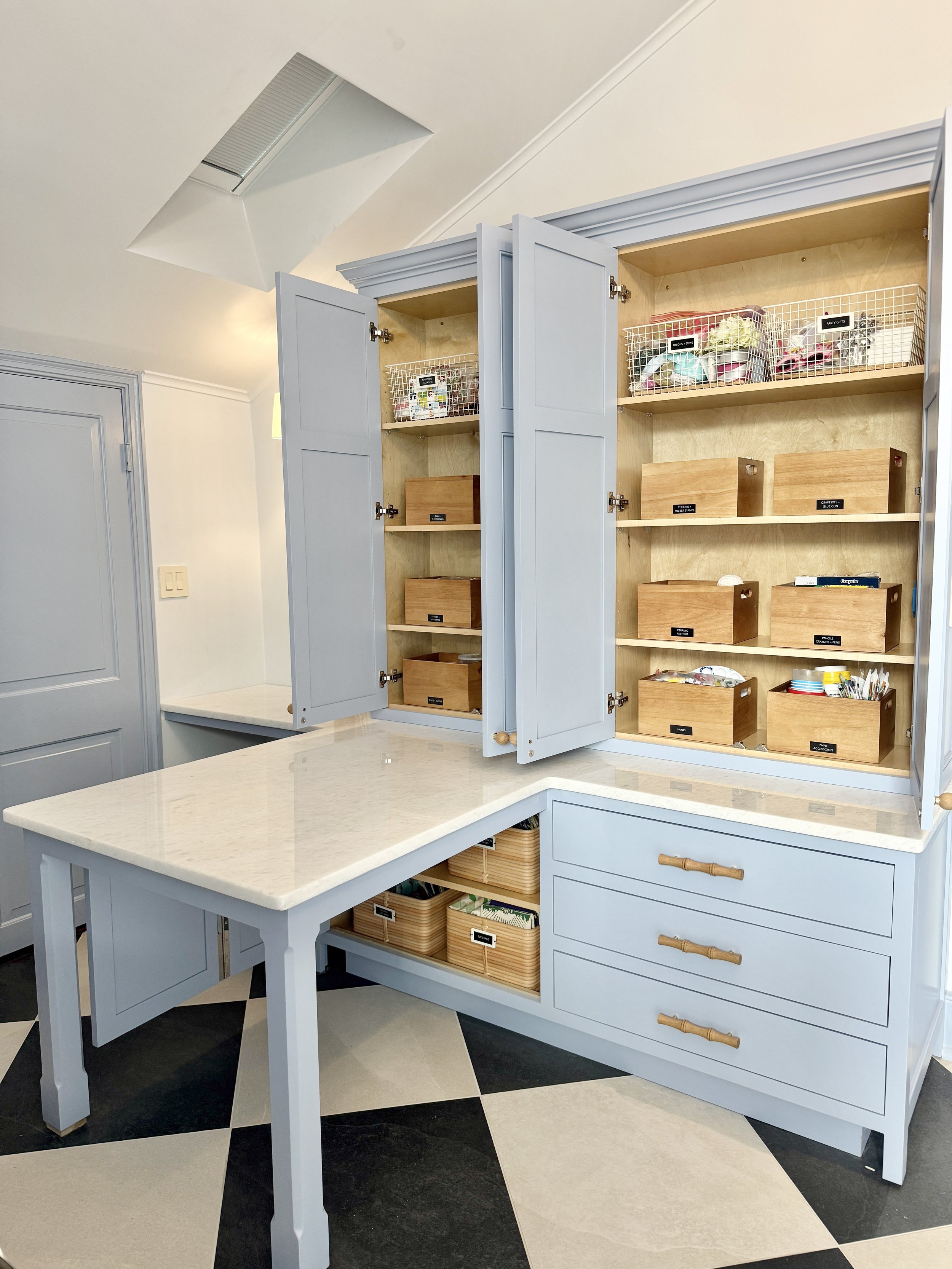 Light blue kitchen cabinet with open doors showing organized wooden boxes and baskets inside. A white marble countertop extends from the cabinet, with black and white checkered flooring below.