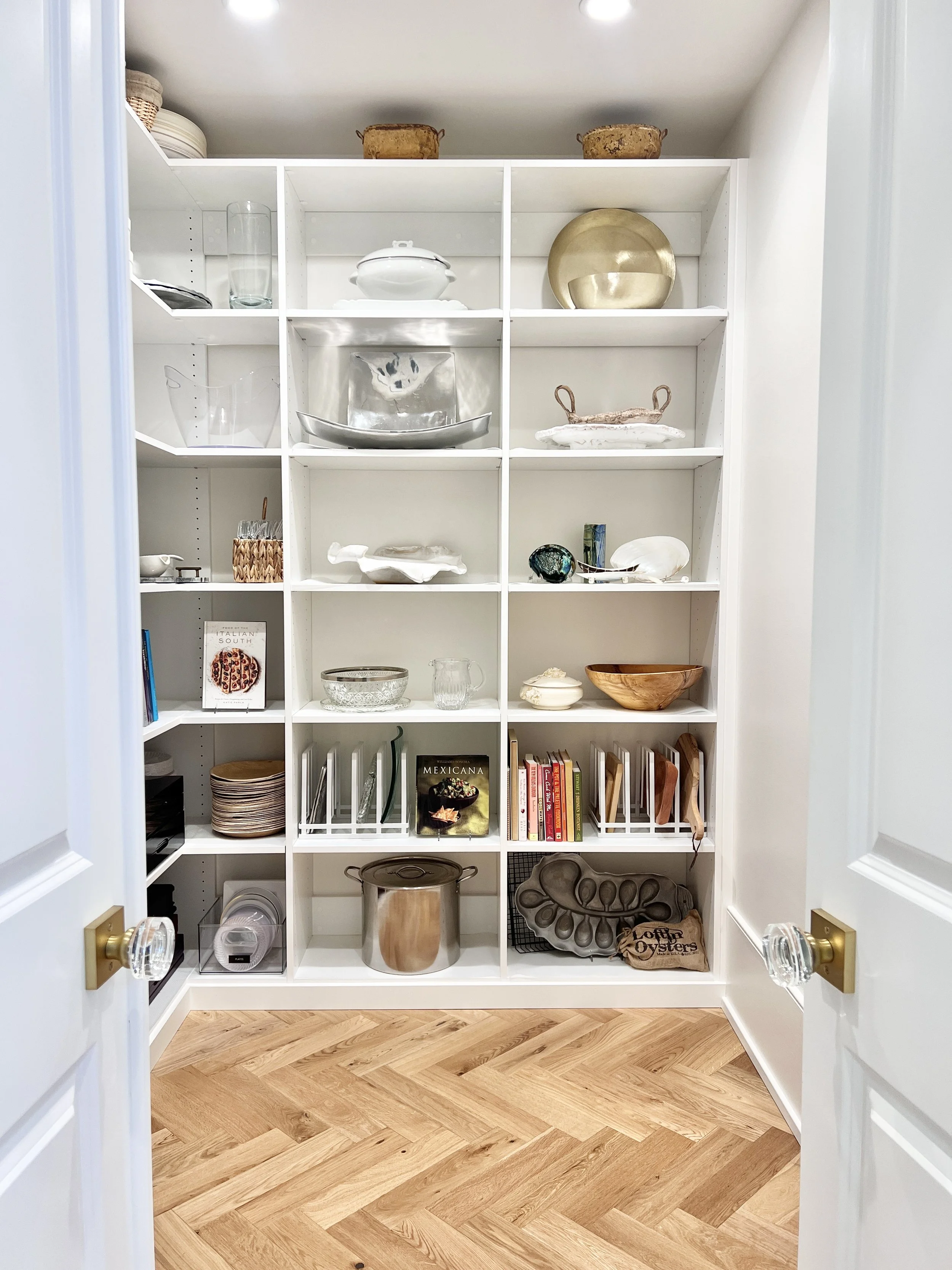 Organized pantry with white shelves featuring curated platters, cookbooks, oyster trays, and cookware, styled by Sorted Professional Organizing.