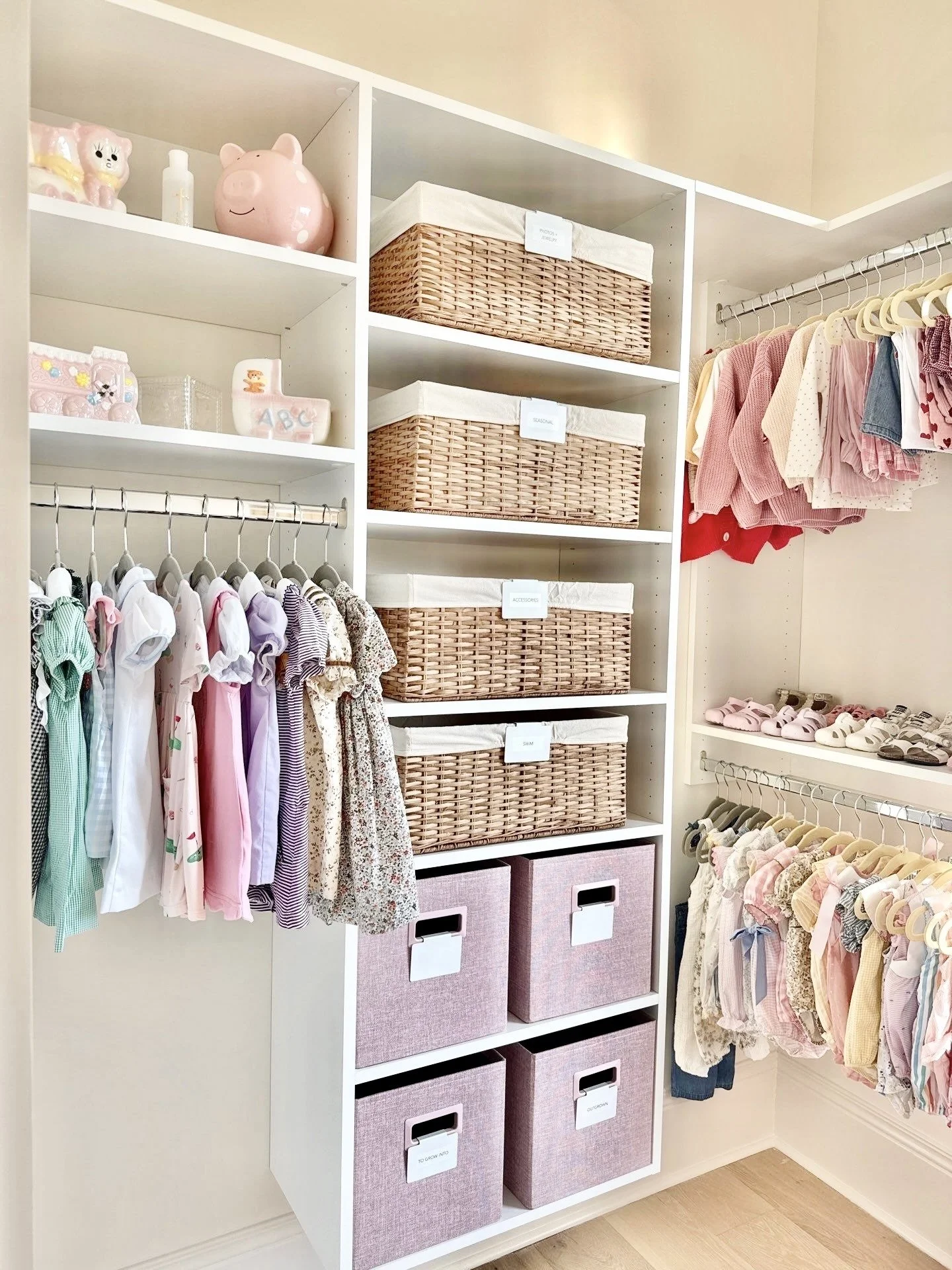 Nursery closet with baby clothes on hangers, woven storage baskets labeled and pink fabric bins, and decorative items including a pink piggy bank and animal figurines.