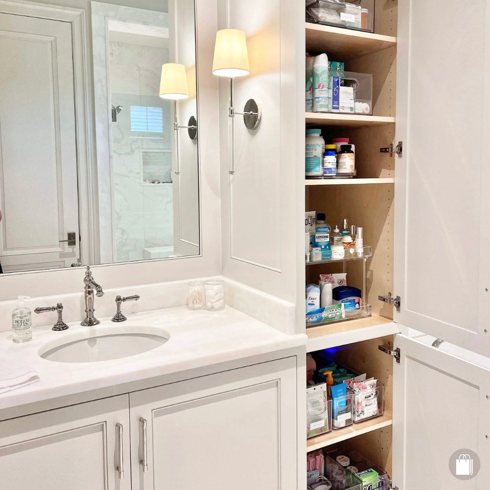 Bathroom with a white vanity, marble countertop, and a mirror. Open bathroom cabinet filled with toiletries and supplies, with a wall-mounted light fixture.