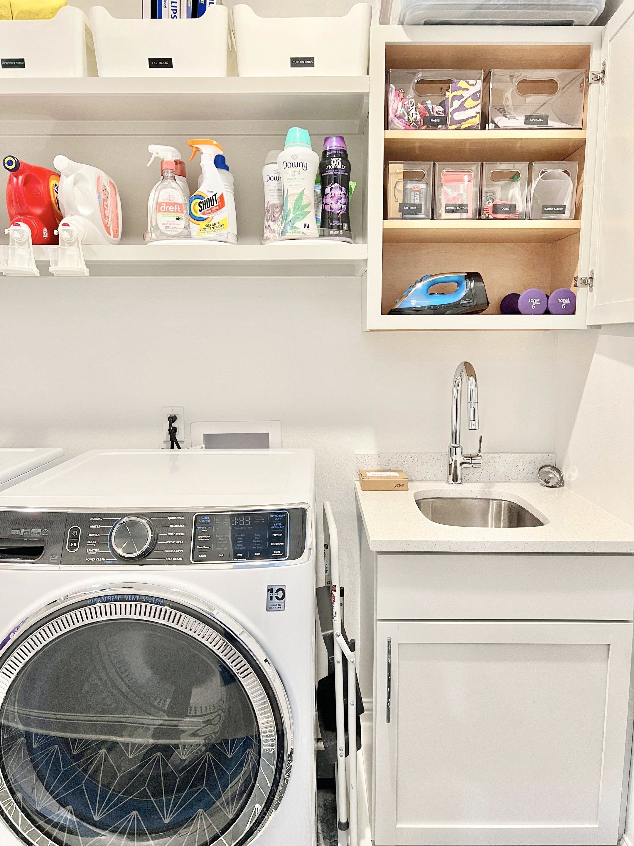 Laundry room with a front-loading washing machine, a utility sink with a cabinet underneath, and wall-mounted shelves holding cleaning supplies, an iron, small containers, and miscellaneous items.
