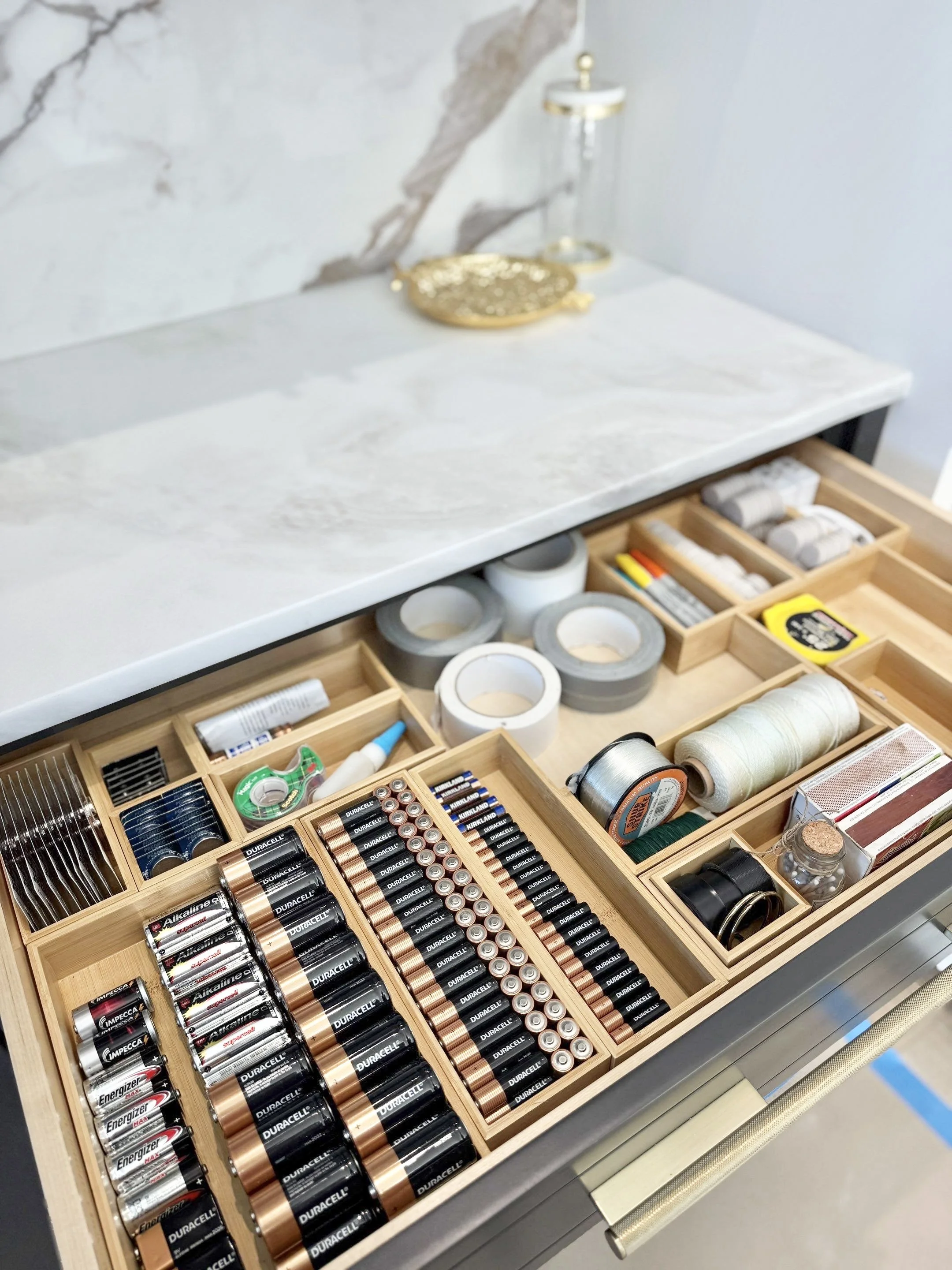 Open utility drawer with bamboo drawer organizers holding various sizes of batteries, duct tape, scotch tape, measuring tape, string, batches, sharpies, and other miscellaneous useful utility drawer items.