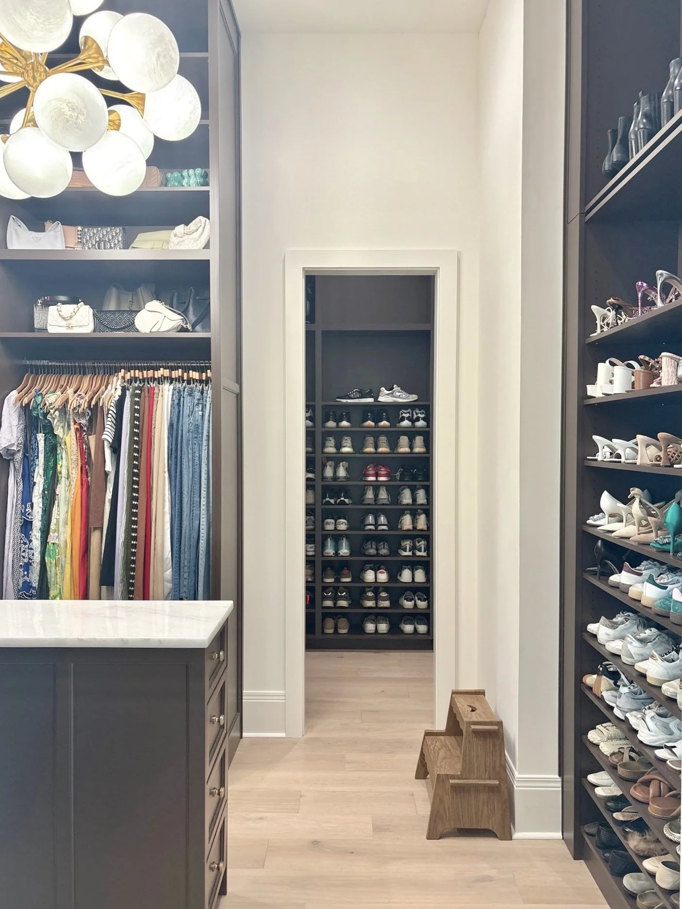 Walk-in closet with shelves of shoes, hanging clothes, handbags, and a small wooden step stool.