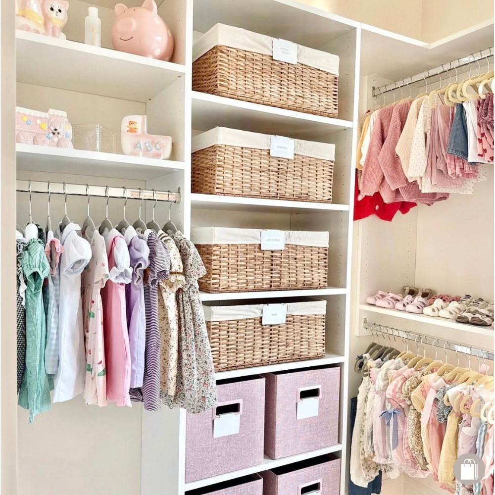 A neatly organized closet shelf with pink, white, and floral children's clothing hanging on a rod, pink and white shoes on a lower shelf, woven baskets on the shelves, and decorative plush toys on top.