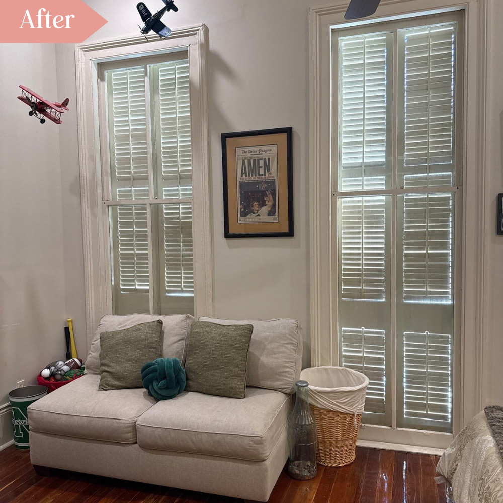 Decluttered room with loveseat, laundry hamper, framed article about the New Orleans Saints reading AMEN, two suspended model airplanes, and closed shutters on tall windows.