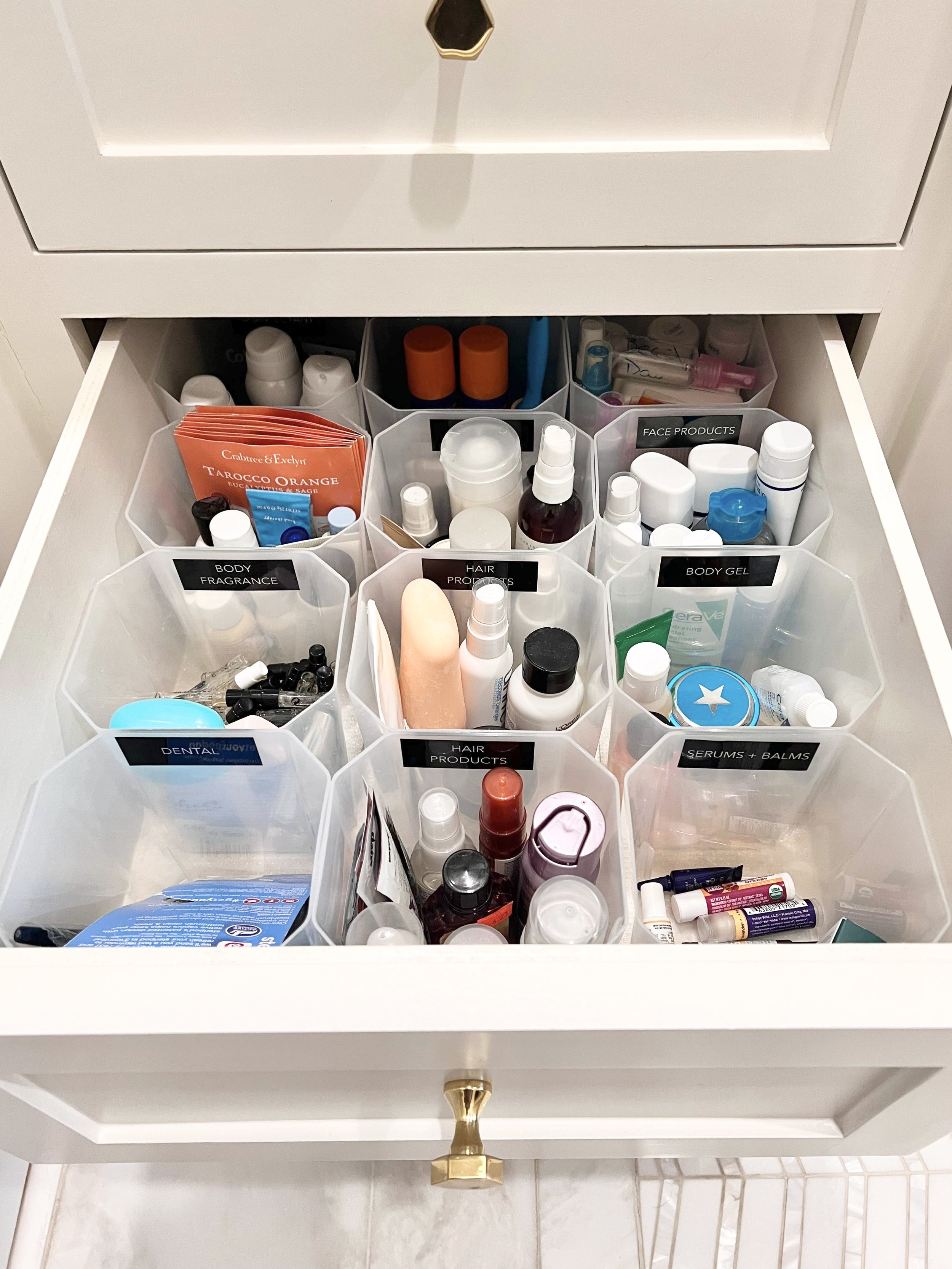 Drawer filled with clear plastic containers holding various personal care and beauty products, each labeled by category such as 'Body Fragrance,' 'Hair Products,' 'Serums + Balms,' and 'Dental.'