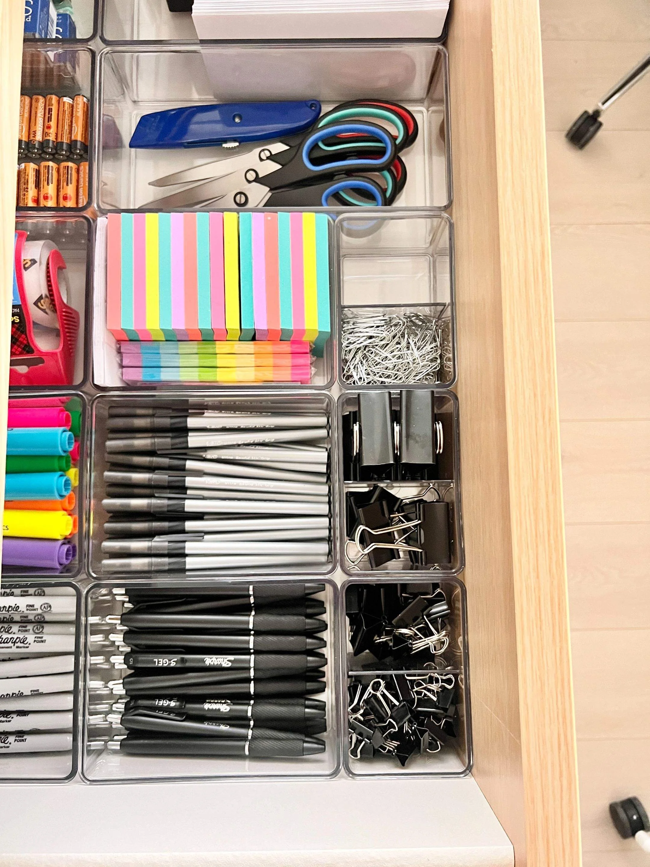 Office desk drawer with acrylic drawer organizers holding a box cutter and scissors, post-it notes, paper clips, binder clips, pins, highlighers, Sharpies, packing tape, and batteries.
