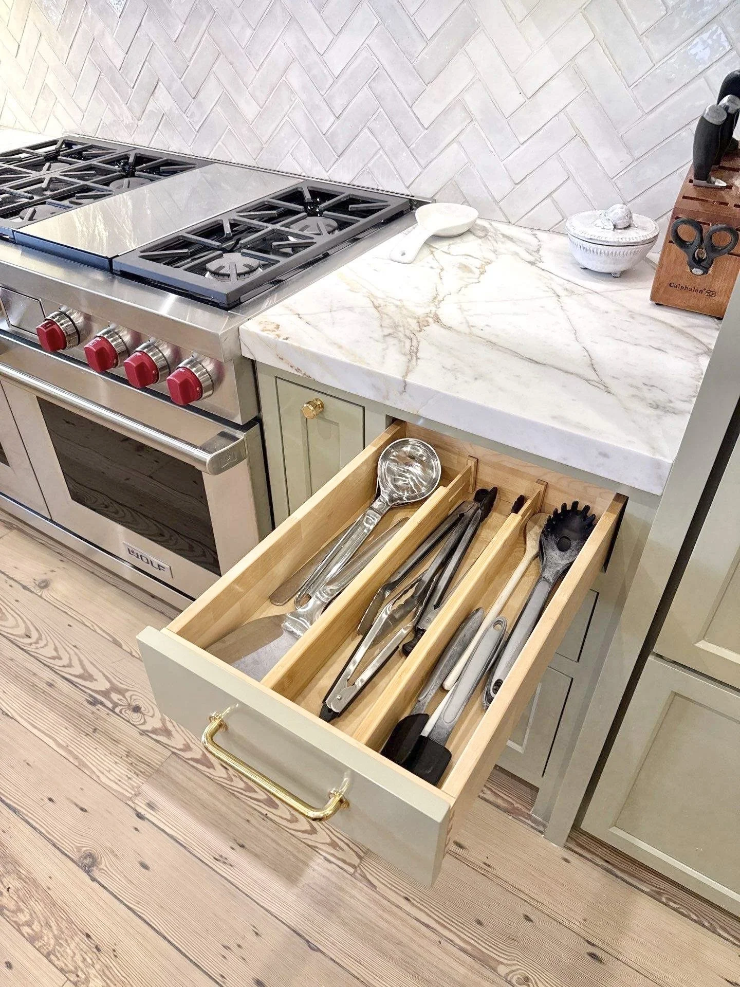 Open itchen drawer with bamboo drawer dividers separating labels, tongs, spatulas, and other kitchen utensils with a gas range oven, white marble countertop with spoon rest and knife block on the countertop.