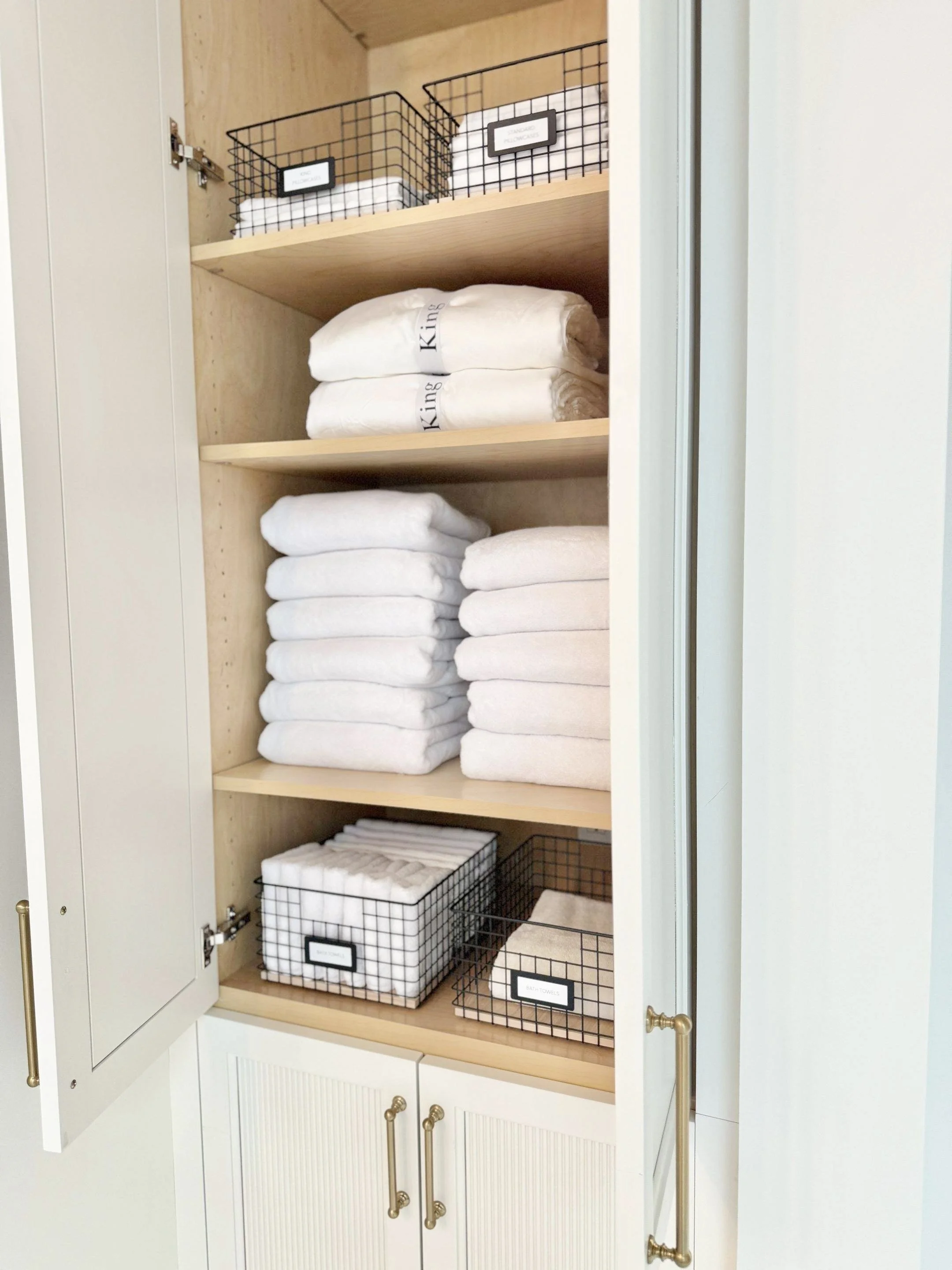 Upper linen cabinet with gold pulls, neatly folded white bath towels, two white folded and labeled king sheet sets, and four black wire grid metal baskets labeled for small hand towels and wash clothes.