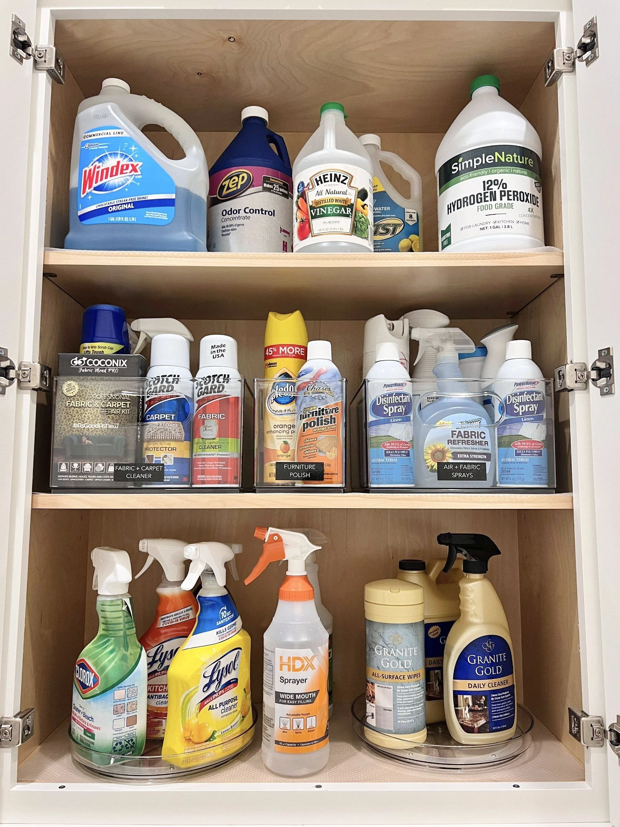 A wooden cabinet filled with cleaning supplies including bleach, vinegar, hydrogen peroxide, fabric and carpet cleaners, furniture polish, disinfectant spray, air and fabric fresheners, spray bottles, and surface wipes.