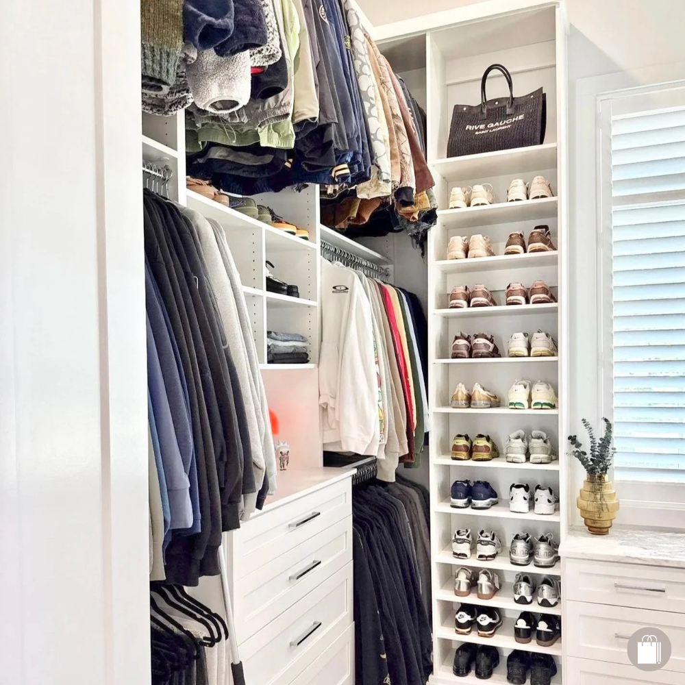 A neatly organized walk-in closet with hanging clothes, shelves of shoes, and a drawer unit. The closet includes shirts, jackets, and pants, with a black handbag on the top shelf and a potted plant on a window ledge.