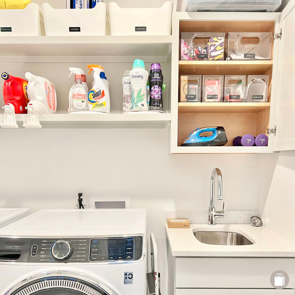 Laundry room with a front-loading washing machine, a utility sink with a cabinet underneath, and wall-mounted shelves holding cleaning supplies, an iron, small containers, and miscellaneous items.