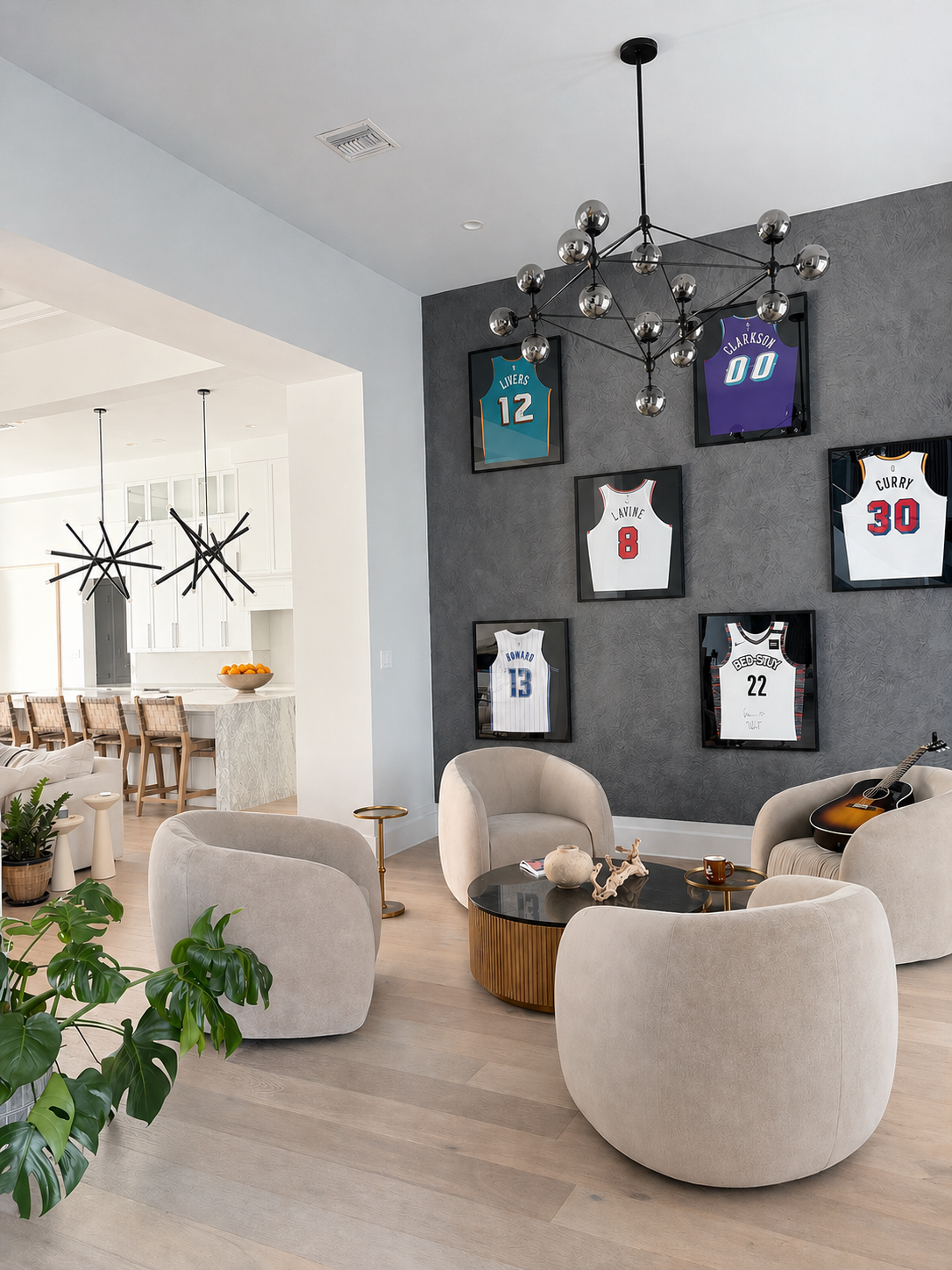 Modern sitting room with four curved lounge chairs around a round coffee table, a dark accent wall displaying framed sports jerseys, and a contemporary chandelier overhead, opening into a bright kitchen with bar stools.