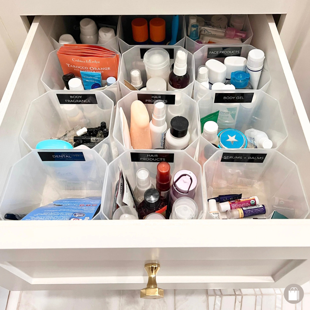 Drawer filled with clear plastic containers holding various personal care and beauty products, each labeled by category such as 'Body Fragrance,' 'Hair Products,' 'Serums + Balms,' and 'Dental.'