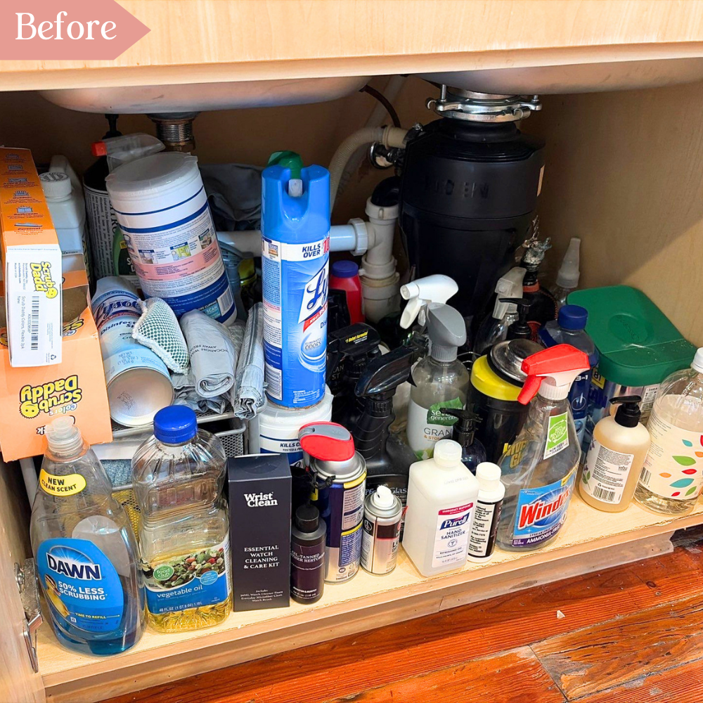 Cluttered under kitchen sink cabinet with stacked cleaning supplies and household products.