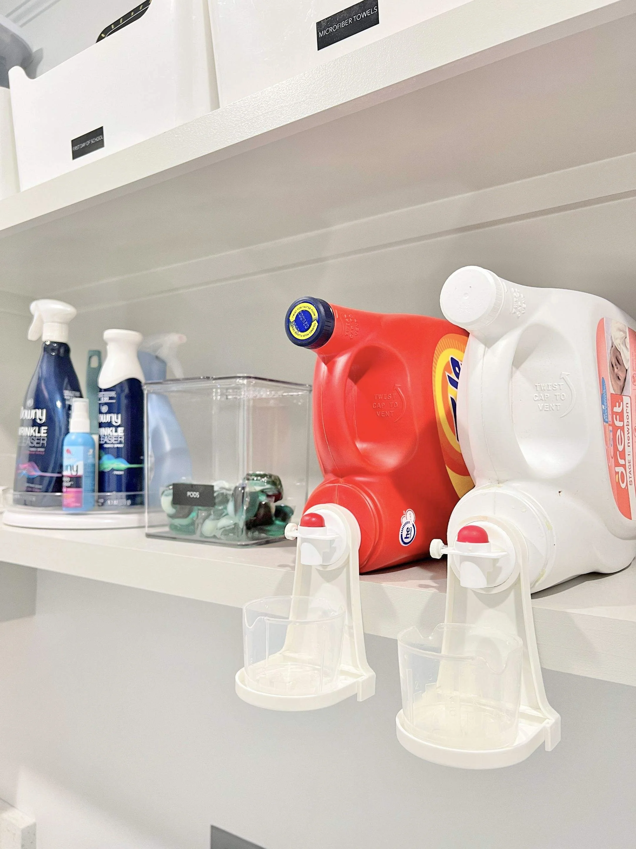Laundry room shelf with detergent bottles, microfiber towels, a container with small miscellaneous items, and two soap dispensers with cups attached.