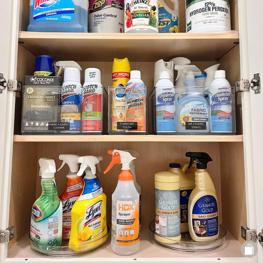 A wooden cabinet filled with cleaning supplies including bleach, vinegar, hydrogen peroxide, fabric and carpet cleaners, furniture polish, disinfectant spray, air and fabric fresheners, spray bottles, and surface wipes.