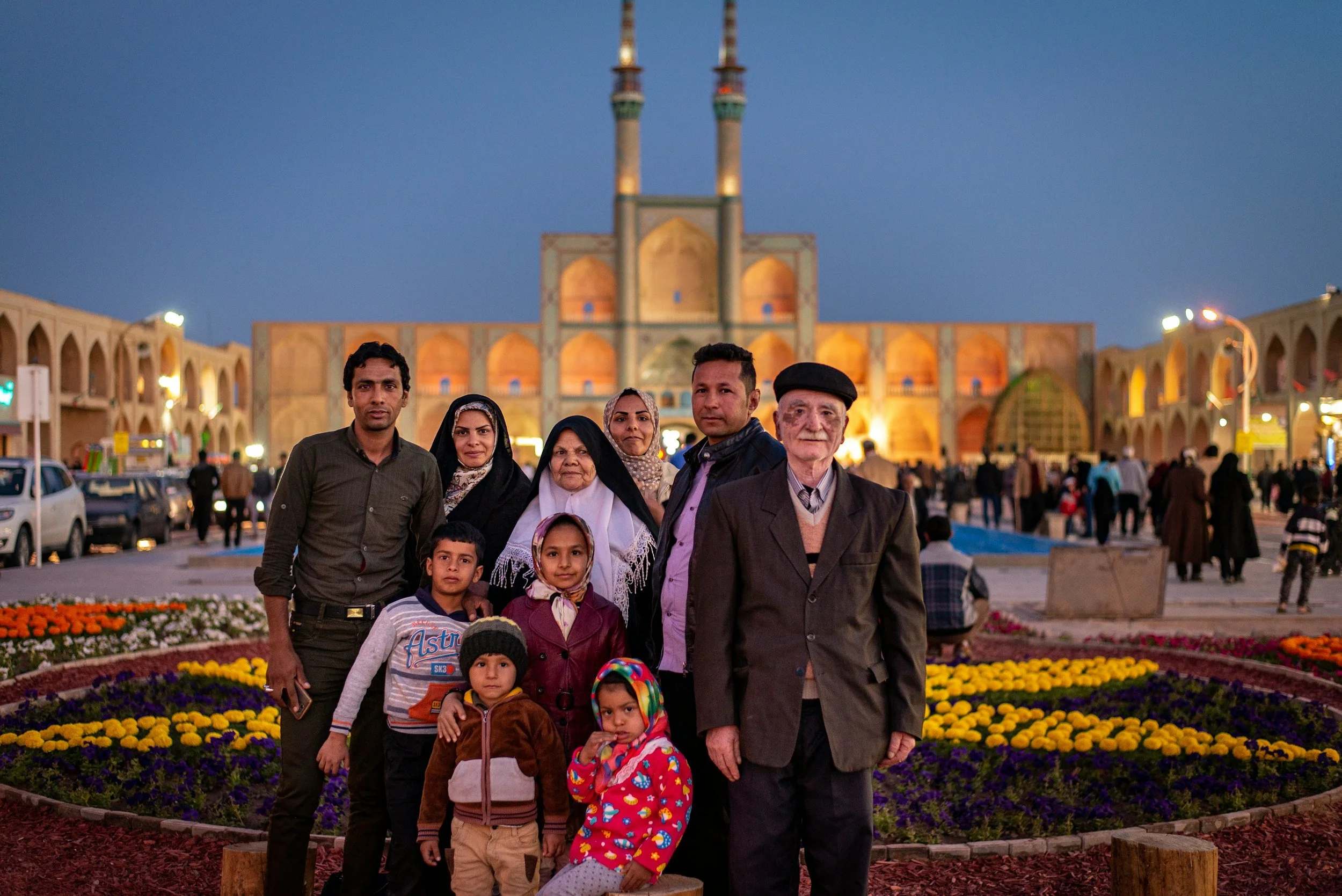 Family of ten posing together in front of an illuminated mosque at dusk with colorful flower beds in the foreground.
