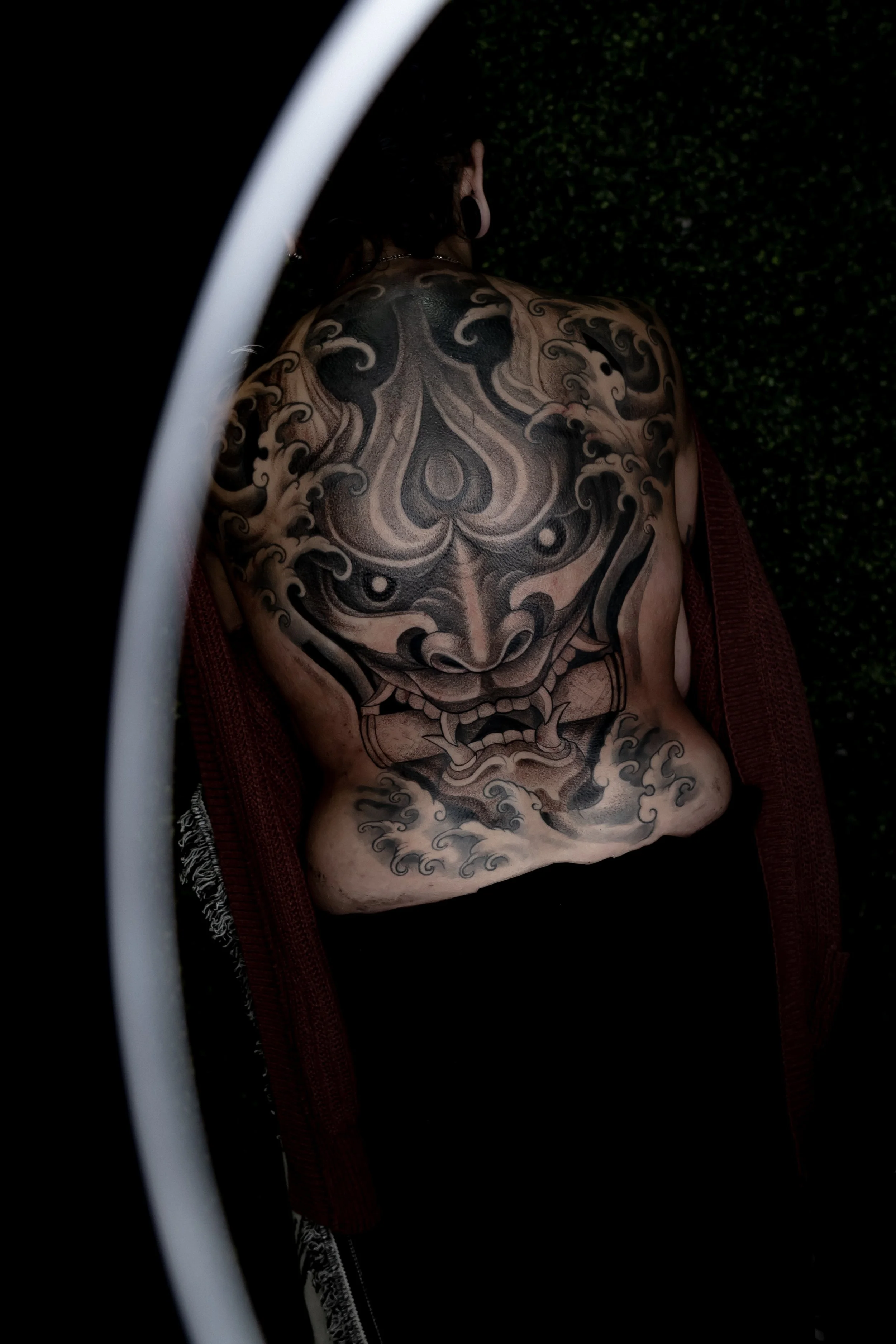 Person with a large tattoo covering their chest and abdomen, partially draped in a red garment, with a dark background and a curved white element in the foreground.