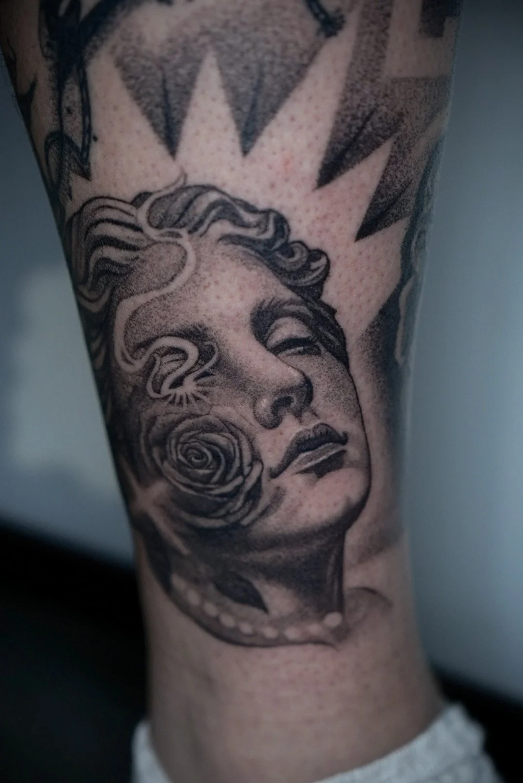 A black and gray tattoo of a woman's face with flowing hair, a rose near her chin, and a starburst pattern above her head.