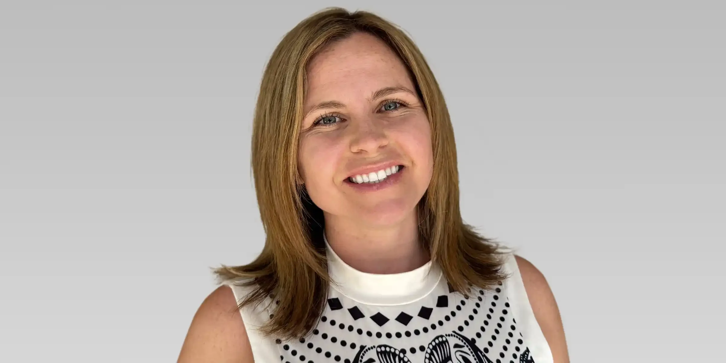 Headshot of a smiling woman with shoulder-length light brown hair, wearing a sleeveless white top with black geometric patterns, against a light gray background.
