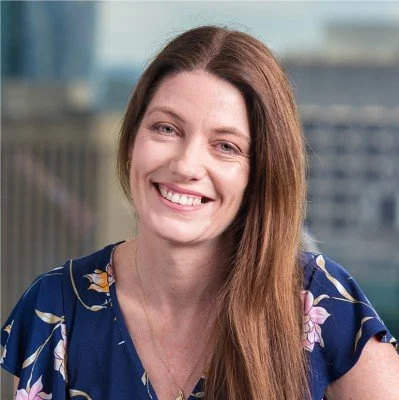 A woman with long red hair smiling at the camera, wearing a blue floral blouse, with a cityscape background.
