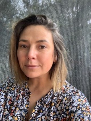 A woman with shoulder-length brown hair and light skin, wearing a floral shirt, standing in front of a window with a screen and trees visible outside.