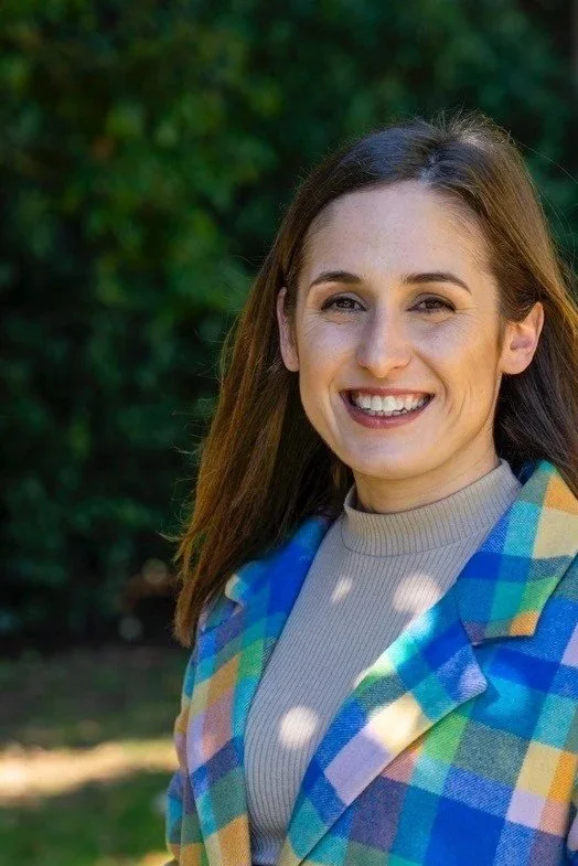 A smiling woman with long brown hair in a colorful checkered jacket standing outdoors with greenery in the background.