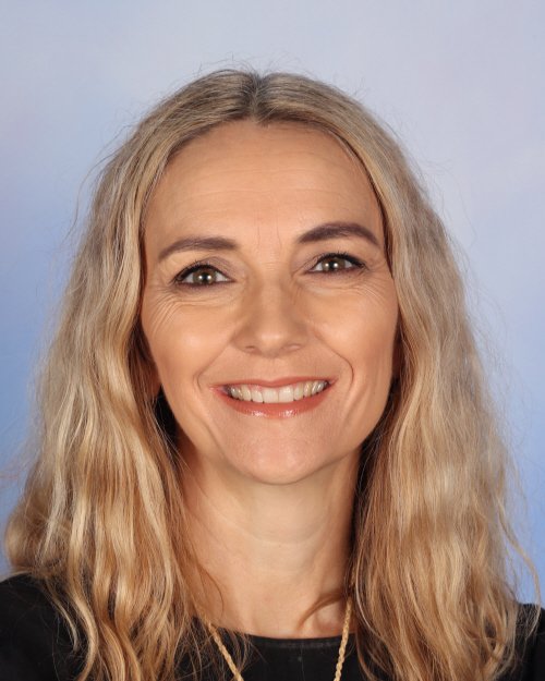 A smiling woman with long, wavy blonde hair, wearing a black top and a necklace, against a light blue background.