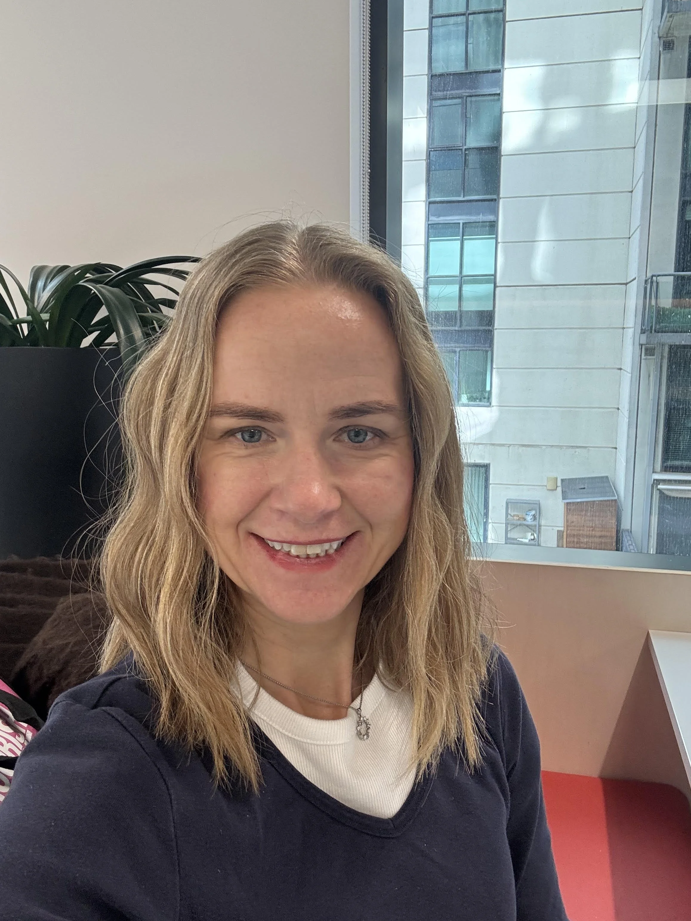 A woman with long blonde hair, smiling, wearing a white top, black cardigan, and a necklace, sitting at a table near a large window with an urban cityscape view behind her.