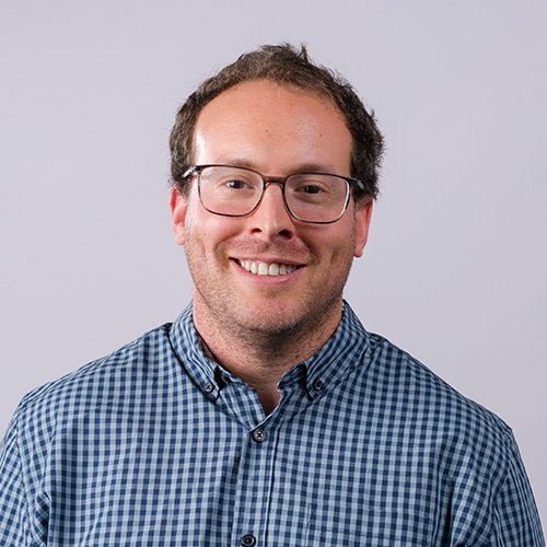 A man with glasses, smiling, in a blue checkered shirt against a plain light gray background.