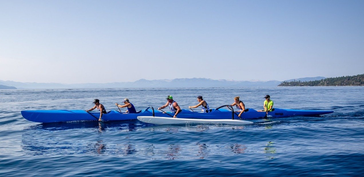 Team paddling a blue outrigger canoe on open water with distant hills and coastline in the background.