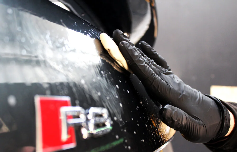 Person wearing black gloves cleaning or applying polish to a black car with a foam applicator.