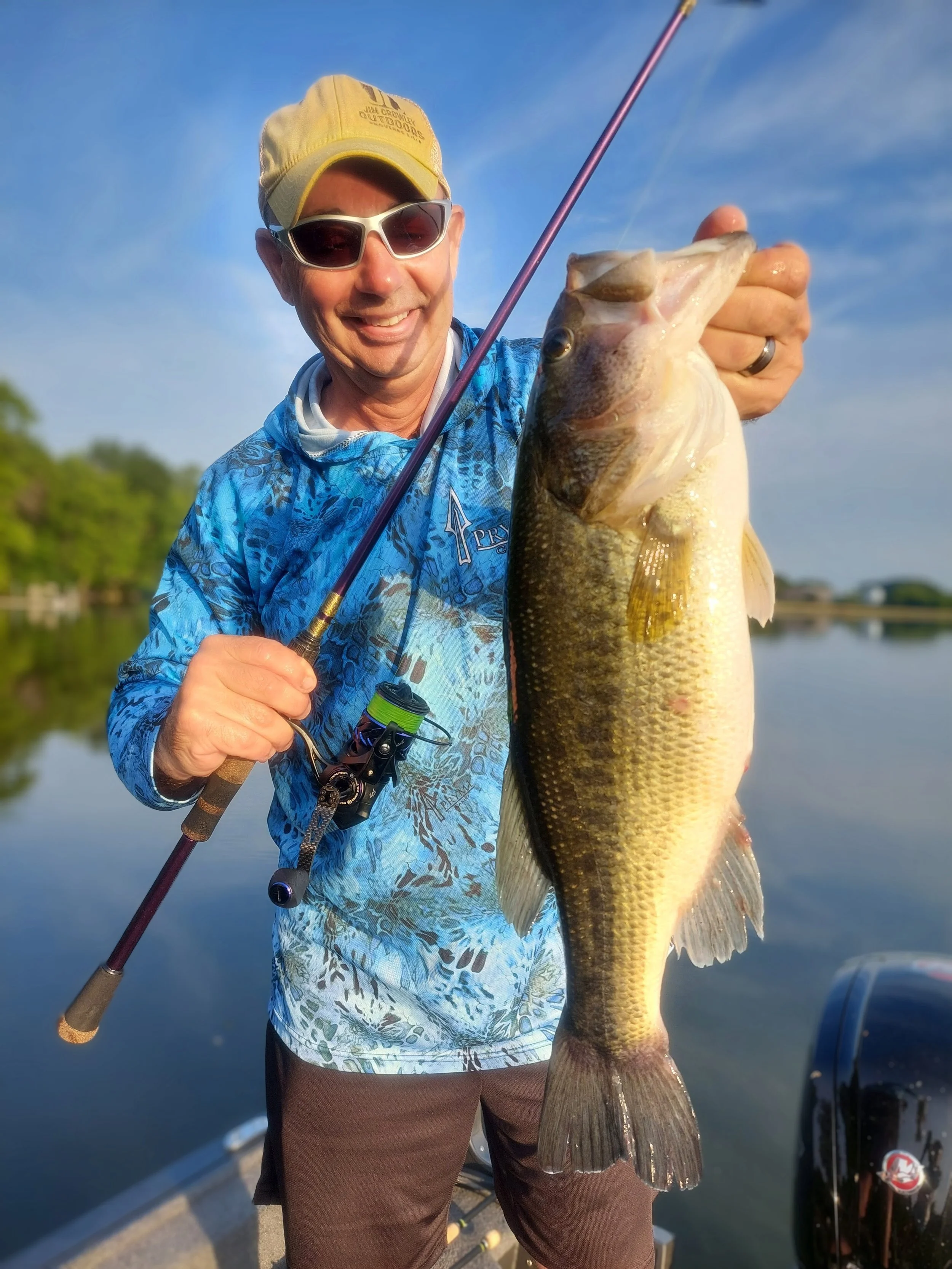 Man wearing a yellow cap, sunglasses, and a blue patterned shirt holding a large bass fish with a fishing rod on a boat in a lake.