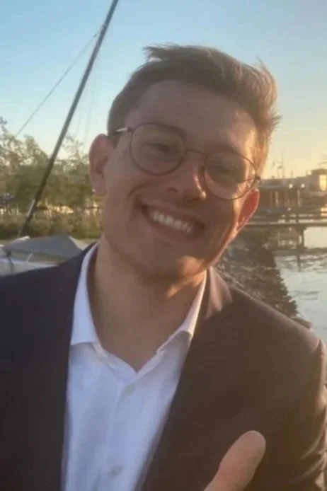 Junger Mann mit Brille und Anzug, lachend, am Wasser bei Sonnenuntergang