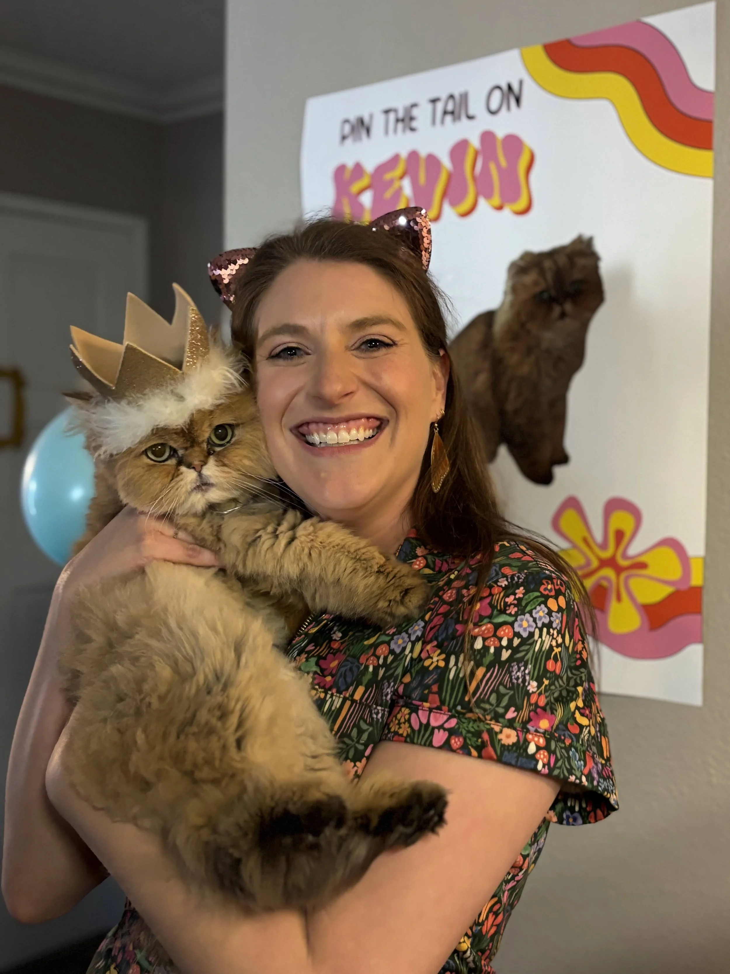 A woman smiling and holding a large fluffy cat wearing a gold paper crown and pink sequin cat ears, standing in front of a colorful birthday banner that reads 'DIY THE TAIL ON.'